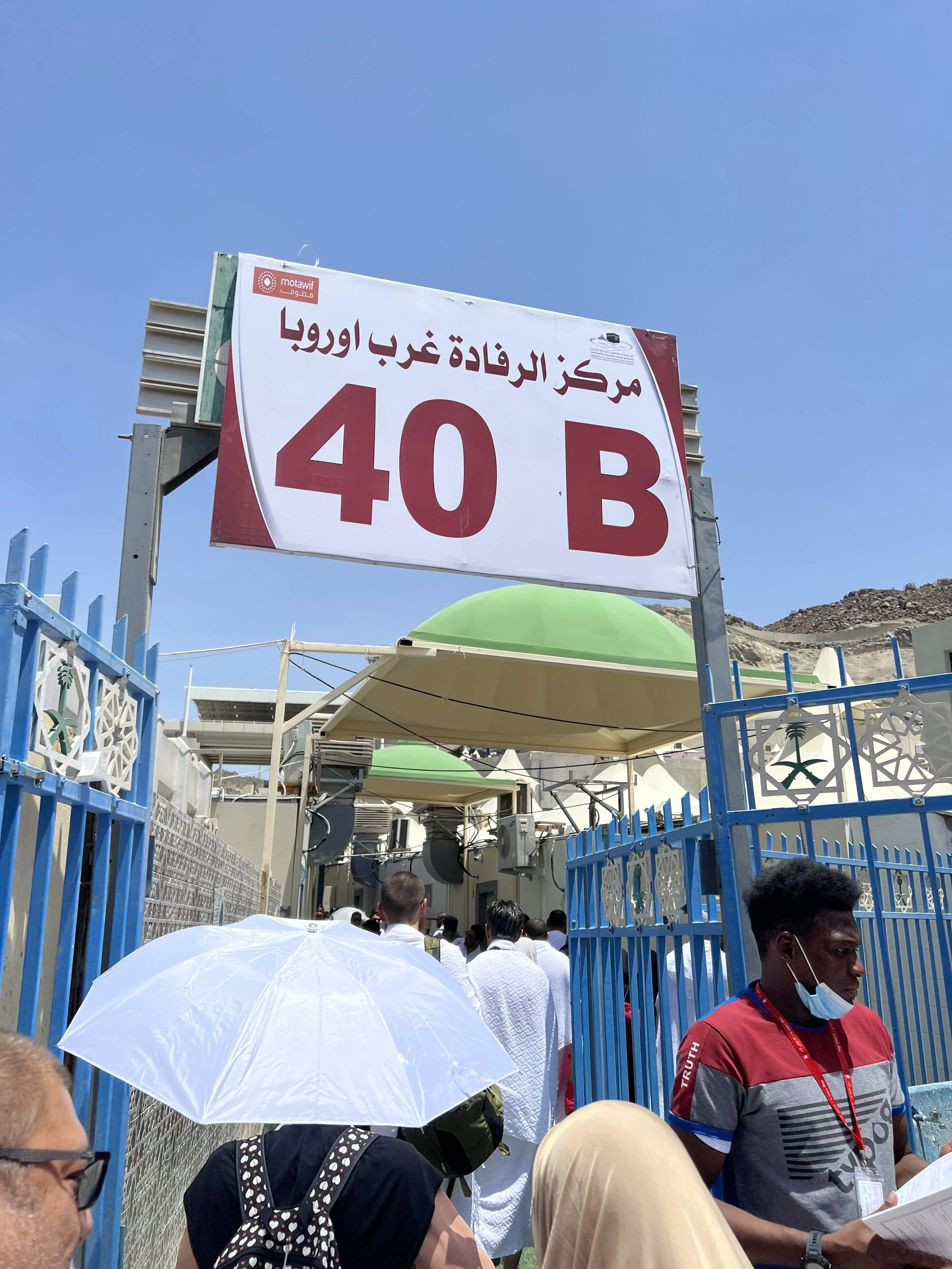 A group of people waiting in line near a blue gate outside a building with a large sign that reads "40 B" in Arabic and English, under a clear blue sky.