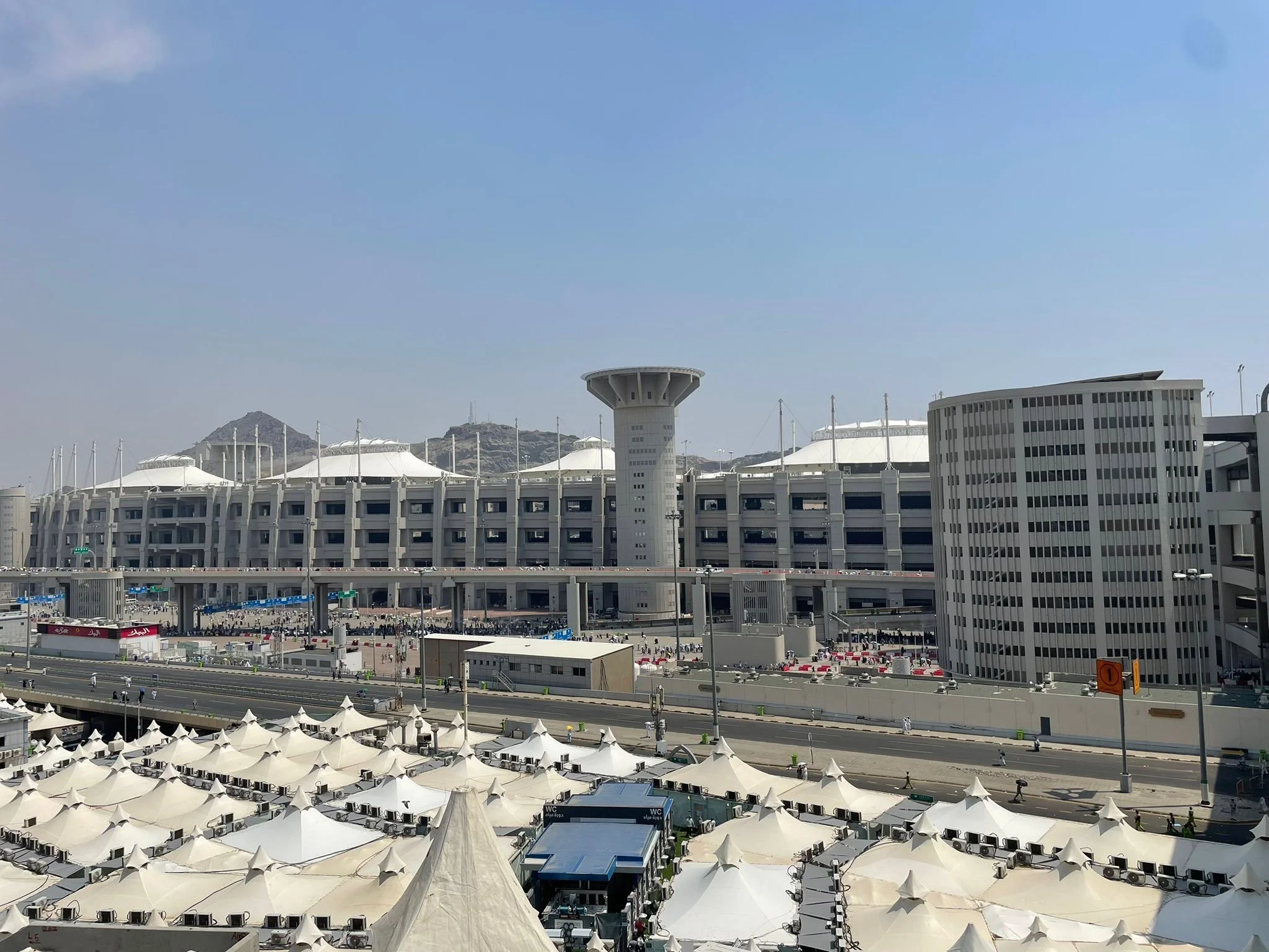 A modern stadium with white tent-like roofs, a central tower, multi-story buildings nearby, and a road with tents and people in the foreground, set against rocky hills.